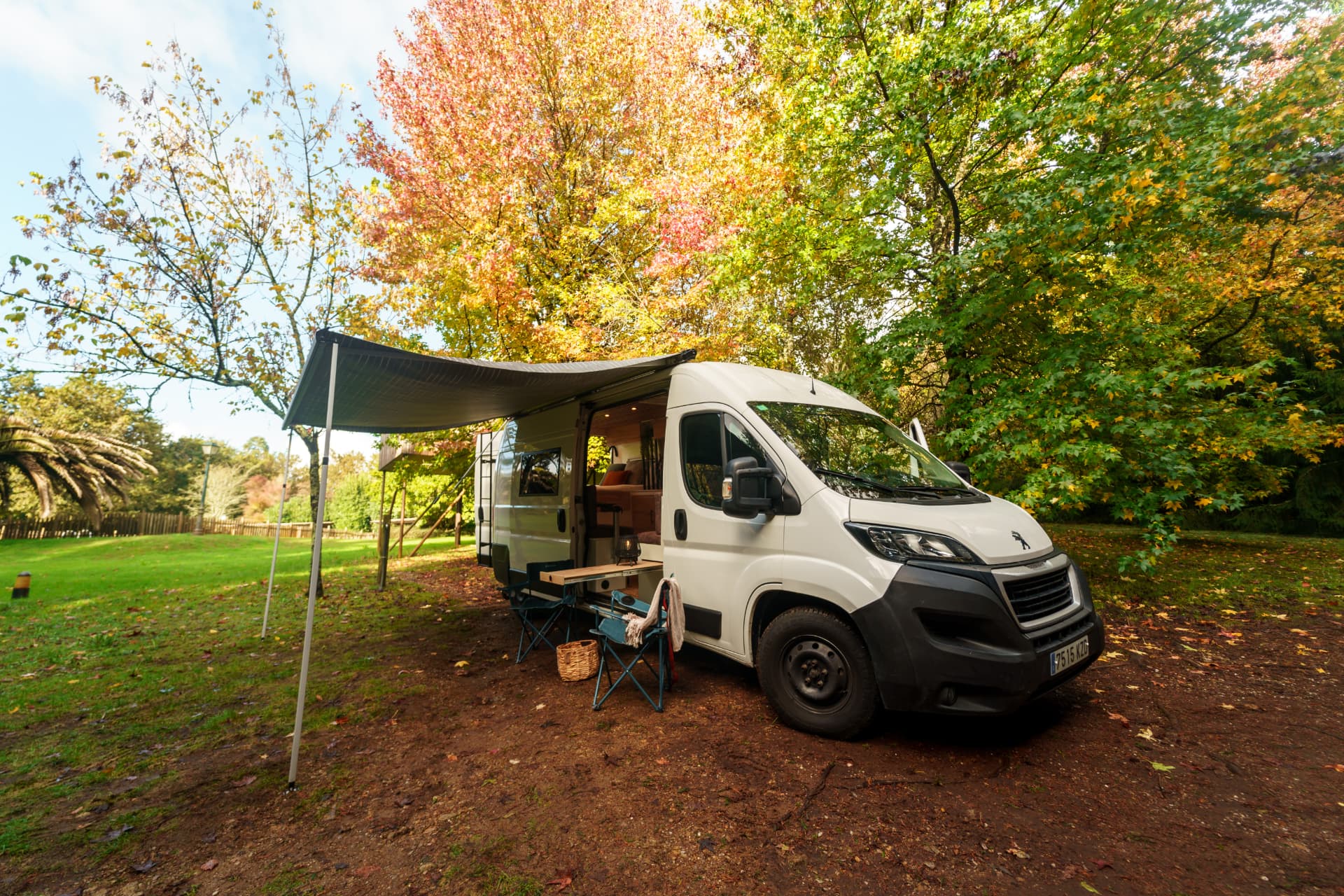 Peugeot Boxer camper blanca con el toldo desplegado, mesa y sillas de camping entre árboles de otoño