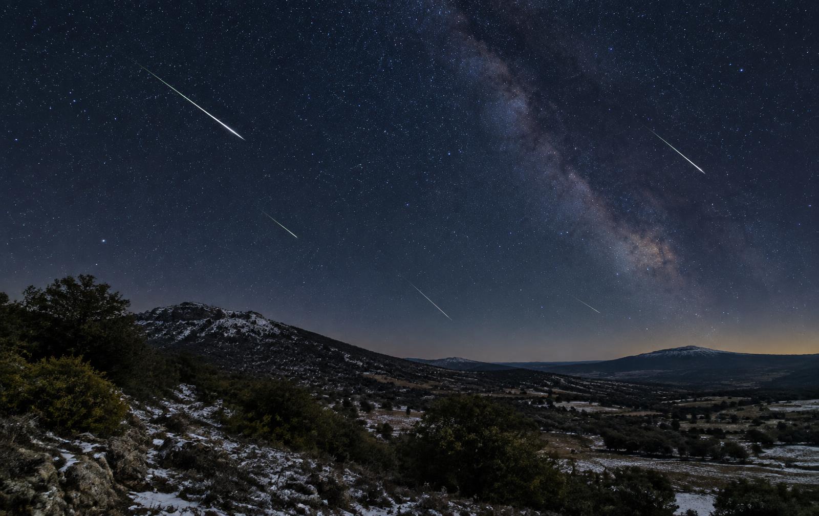 Cielo nocturno estrellado con trazos de meteoros sobre un paisaje de montaña